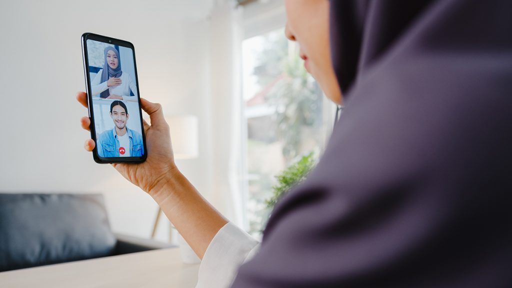 Young Asia muslim student using smart phone talk to peer by videochat brainstorm online meeting while remotely work from home at living room. Social distancing, quarantine for corona virus
