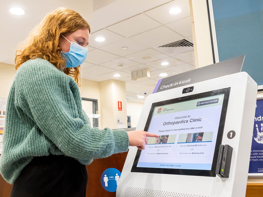 Young woman with mask interacting with digital kiosk in hospital